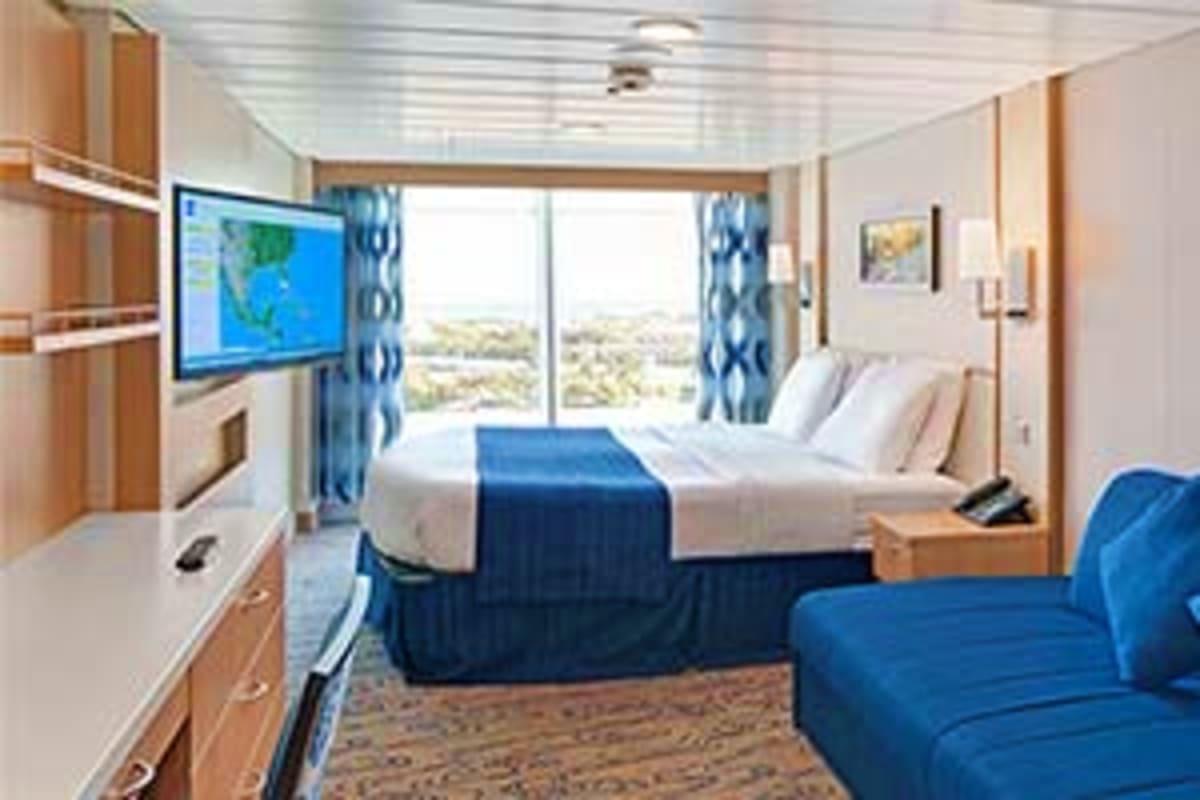 Modern cruise ship stateroom with two beds, blue armchair, flat-screen TV showing map, and ocean view through large balcony w