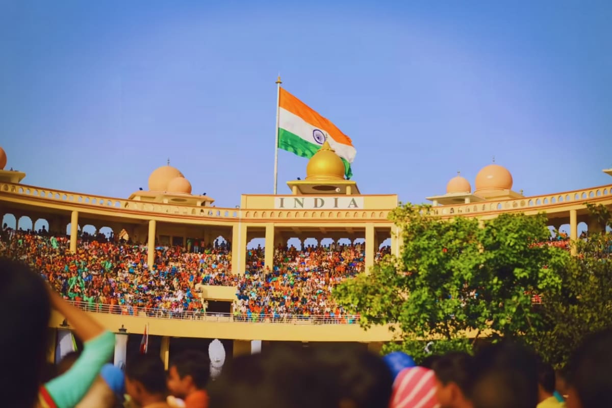 Indian flag flying above a crowded stadium with golden domes and "INDIA" text, spectators in orange clothing watching a borde