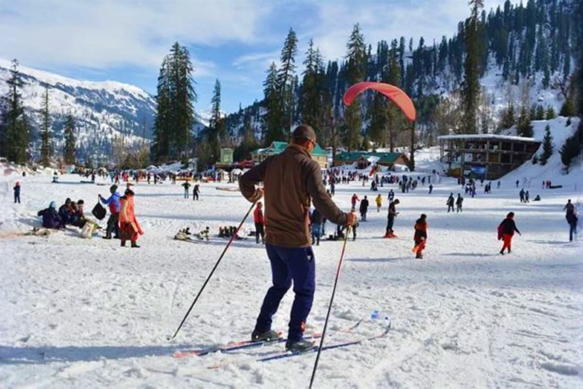 Paraglider in red parachute flying over snow-covered Solang Valley with frozen lake, snow-capped mountains, pine forests, and