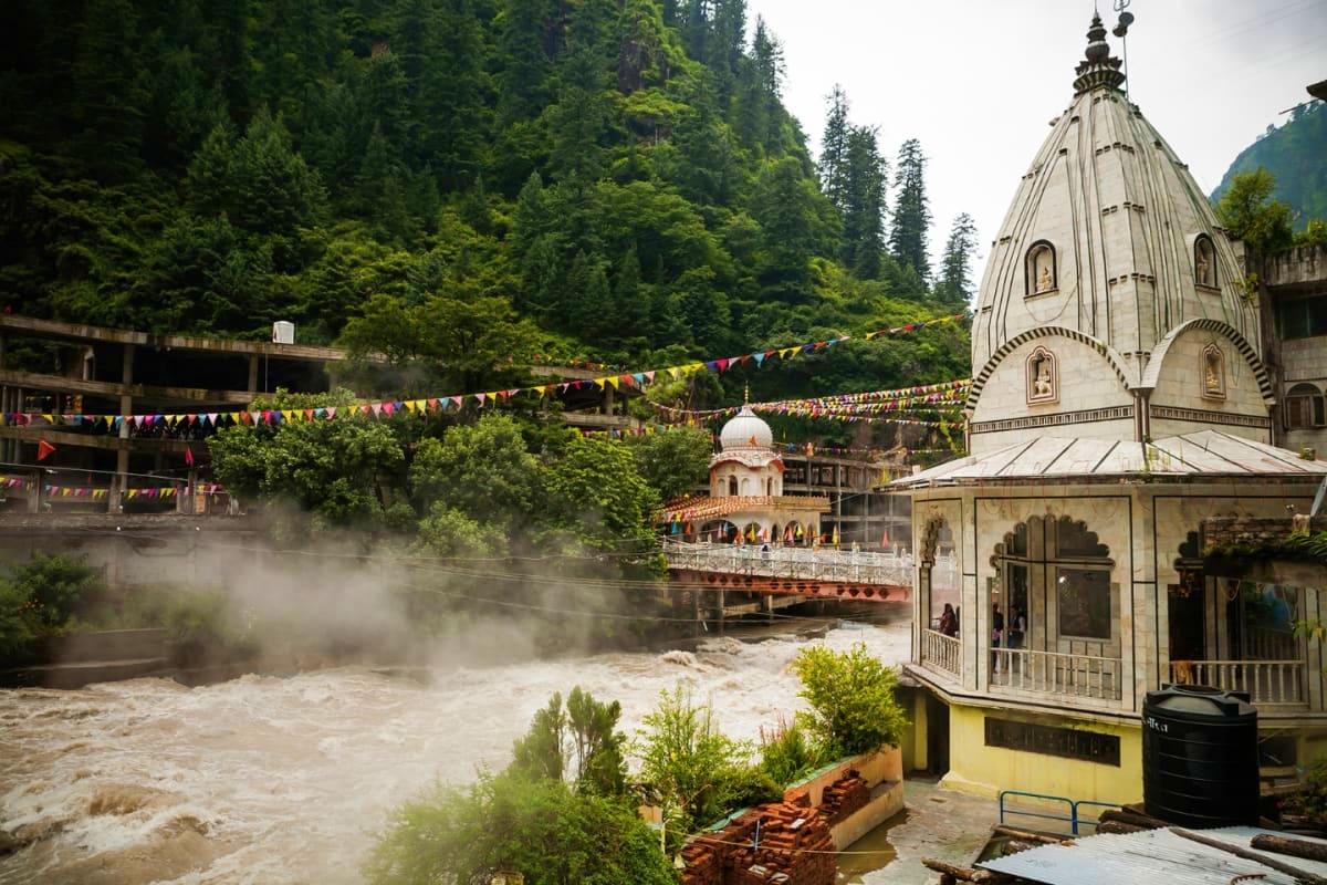 Hindu temple with ornate dome beside turbulent river in forested mountain valley with colorful prayer flags strung overhead
