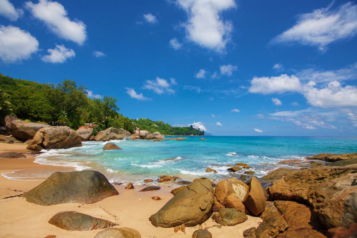 Tropical beach in Seychelles with turquoise water, golden sand, granite boulders, lush green vegetation, and blue sky with wh