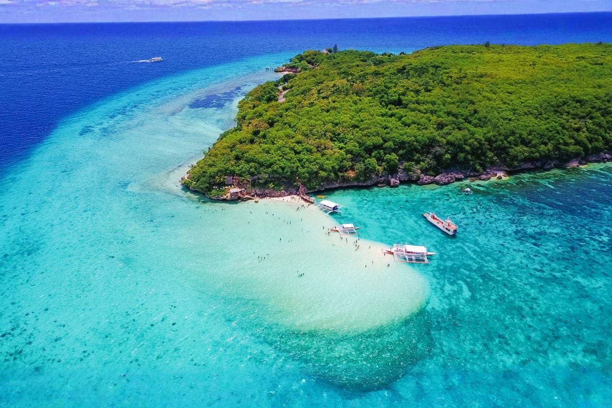 Aerial view of a tropical island with turquoise waters, white sand beach, dense green vegetation, and tourist boats anchored 