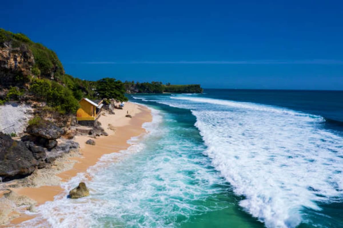 Tropical beach in Bali with white sand, turquoise waves, limestone cliffs, yellow beach hut, and forested coastline under cle