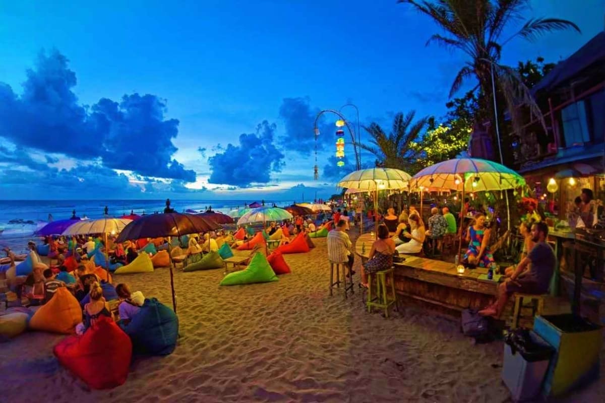 Bali Premium Island Retreat Twilight beach scene in Bali with colorful bean bags, umbrellas, palm trees, and beachfront bars with crowds of people