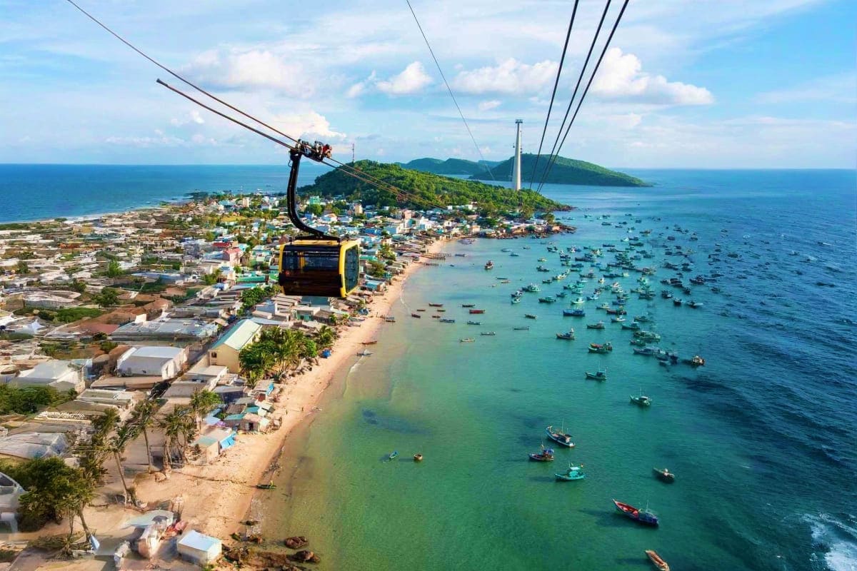 Halong Waves and Phu Quoc Days Aerial view of a yellow cable car cabin over Phu Quoc island beach with turquoise water, fishing boats, and coastal town belo