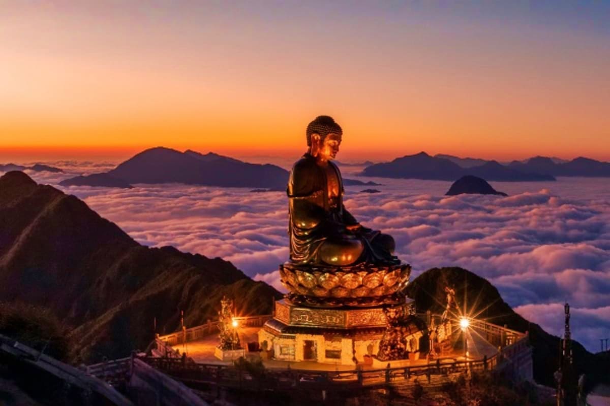Golden Buddha statue illuminated at sunset on mountainside above sea of clouds with distant peaks