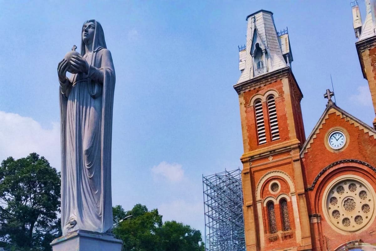 Stone statue of Virgin Mary holding a cross beside red brick cathedral with bell tower and clock against blue sky