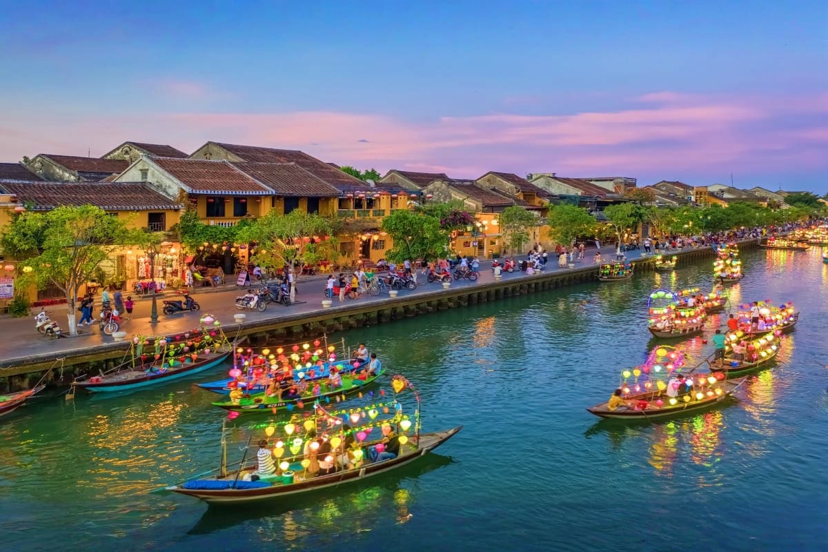 Hoi An ancient town at dusk with illuminated lantern boats on Thu Bon River and yellow colonial buildings along the waterfron