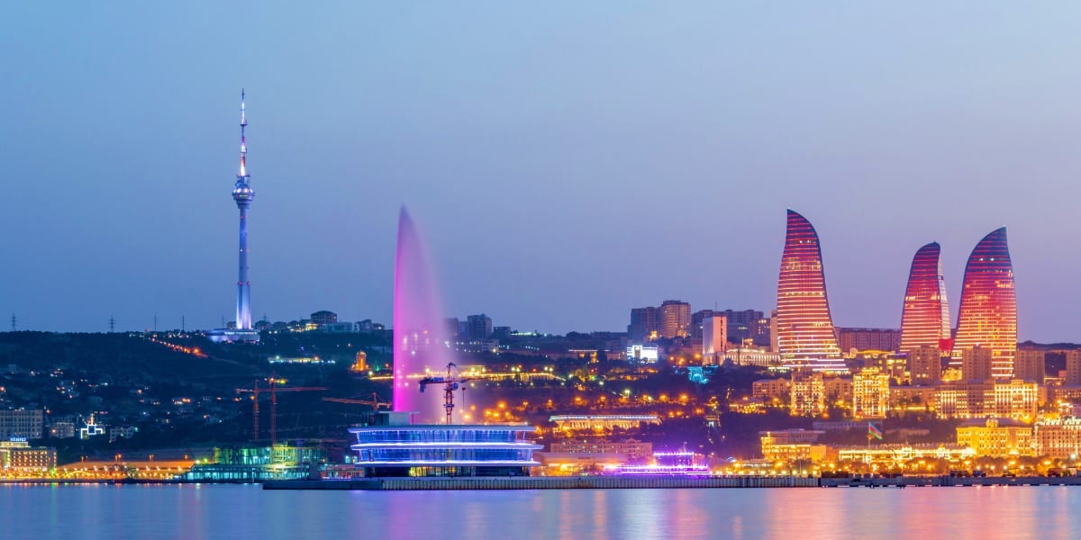 Baku cityscape at dusk with illuminated Flame Towers, glowing fountain, and TV tower reflected in Caspian Sea water