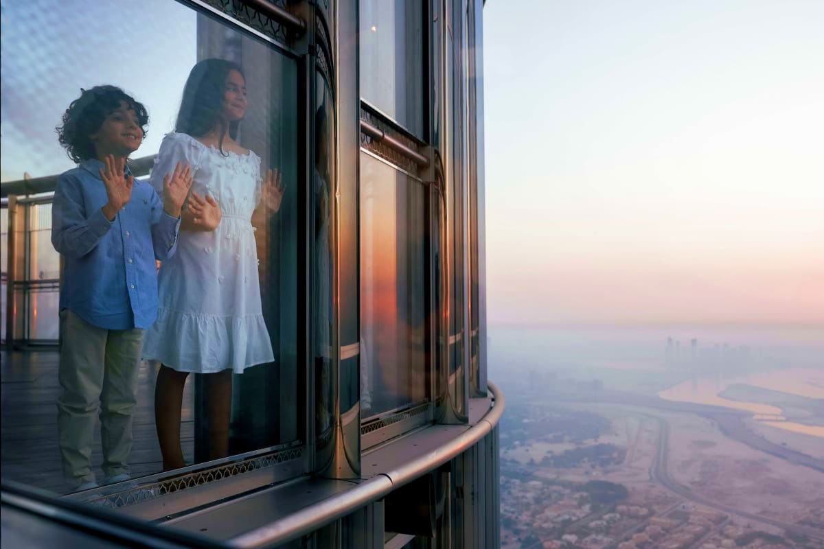 Two children looking out from a high-rise window at sunset over a cityscape below from the Burj Khalifa observation deck