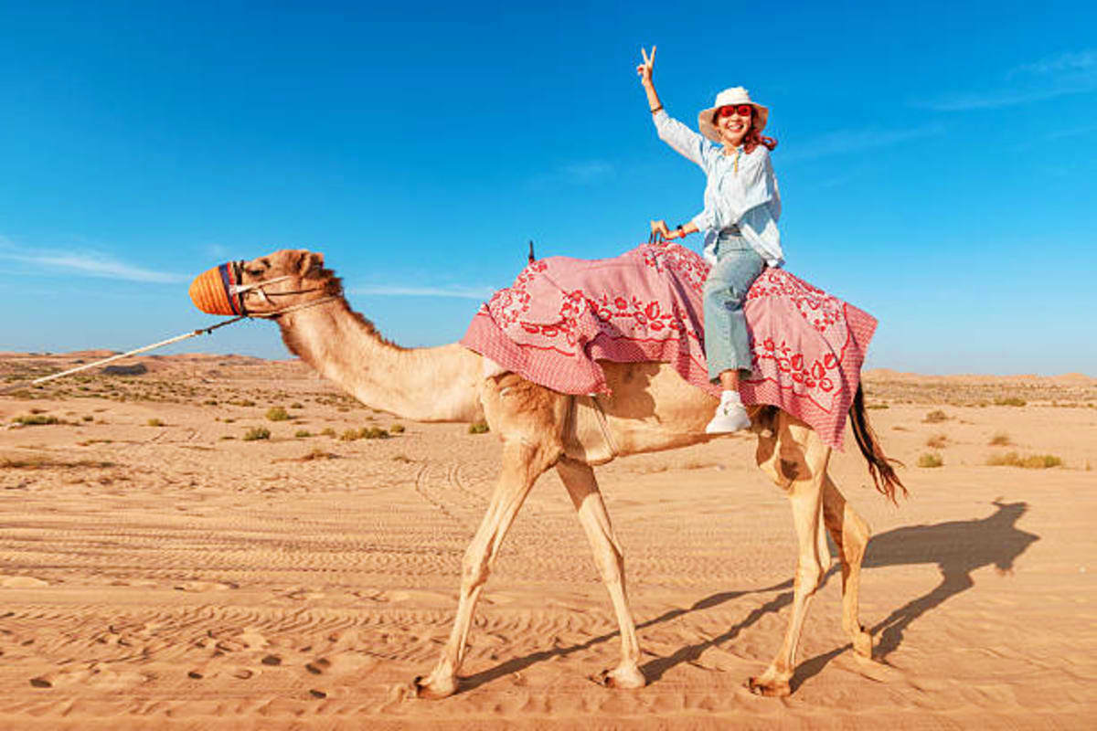 Camel Riding Woman riding a camel in the desert while making a peace sign, wearing a white hat and red decorative saddle blanket