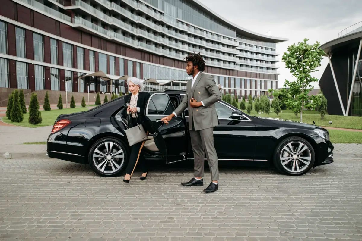 Professional driver assisting senior woman exiting luxury black sedan outside modern high-rise building in urban setting