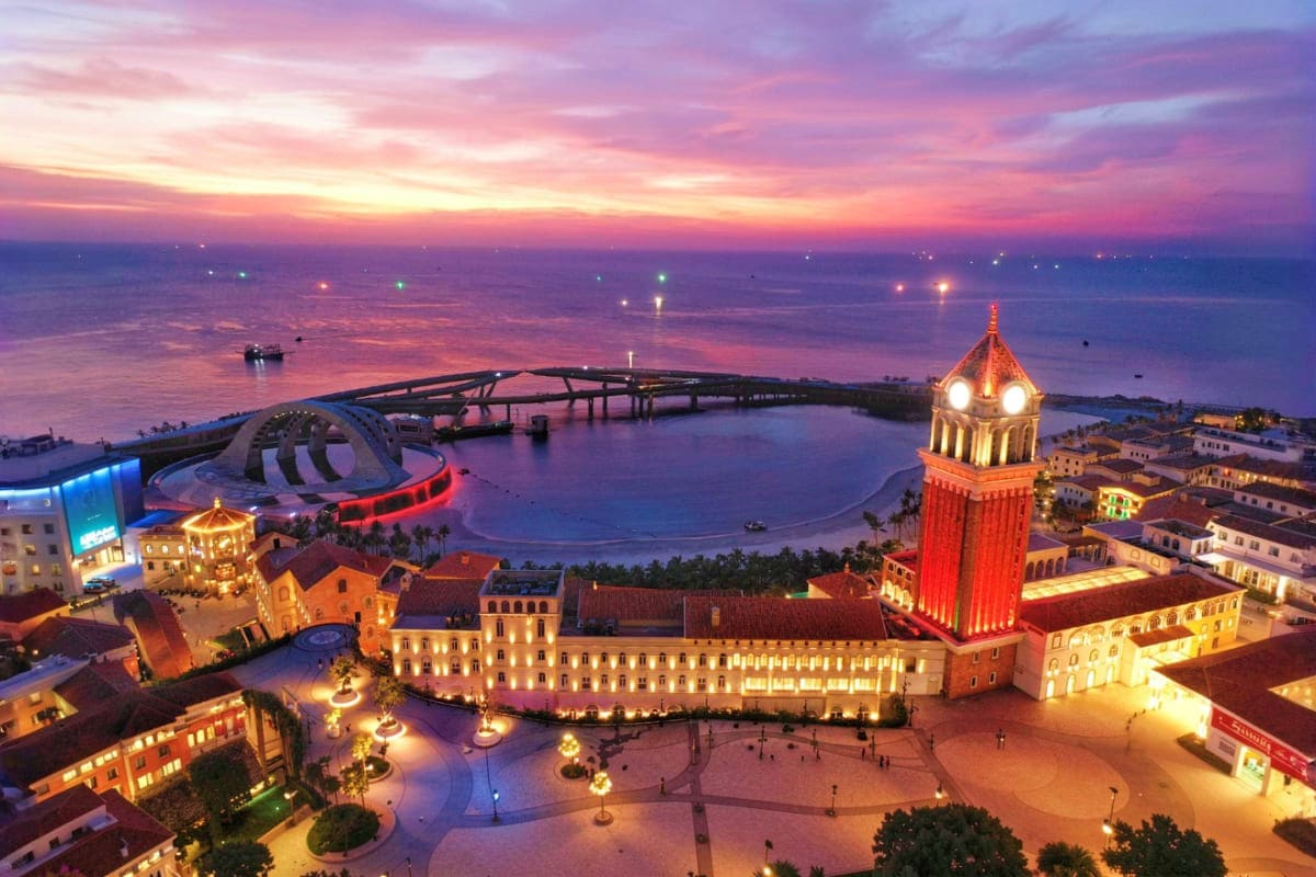 Aerial sunset view of waterfront resort with illuminated tower, domed structures, curved bridge, and bay with boats at dusk