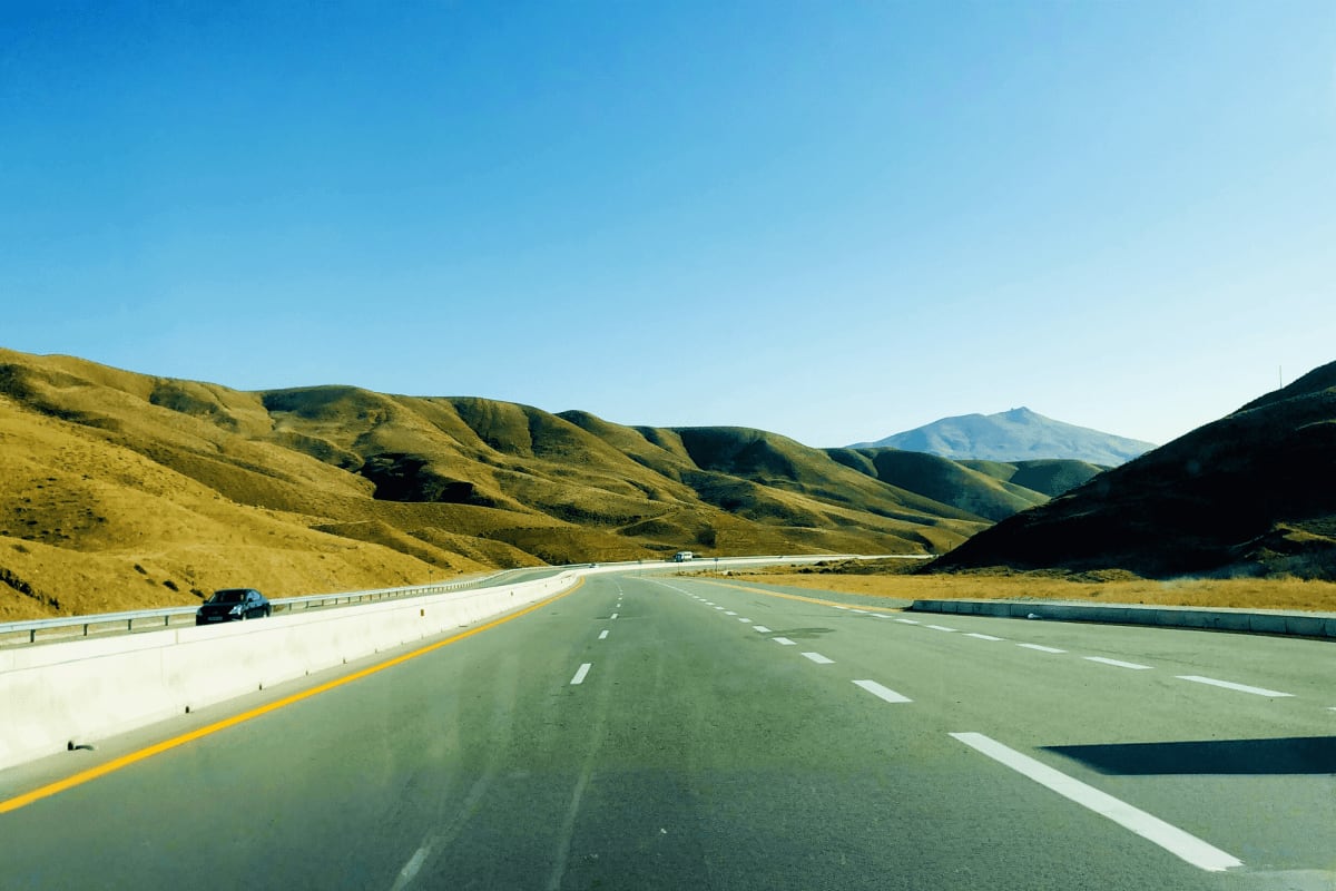 Baku to Shahdag Tour Multi-lane highway stretching through rolling green mountains with snow-capped peak in distance, clear blue sky