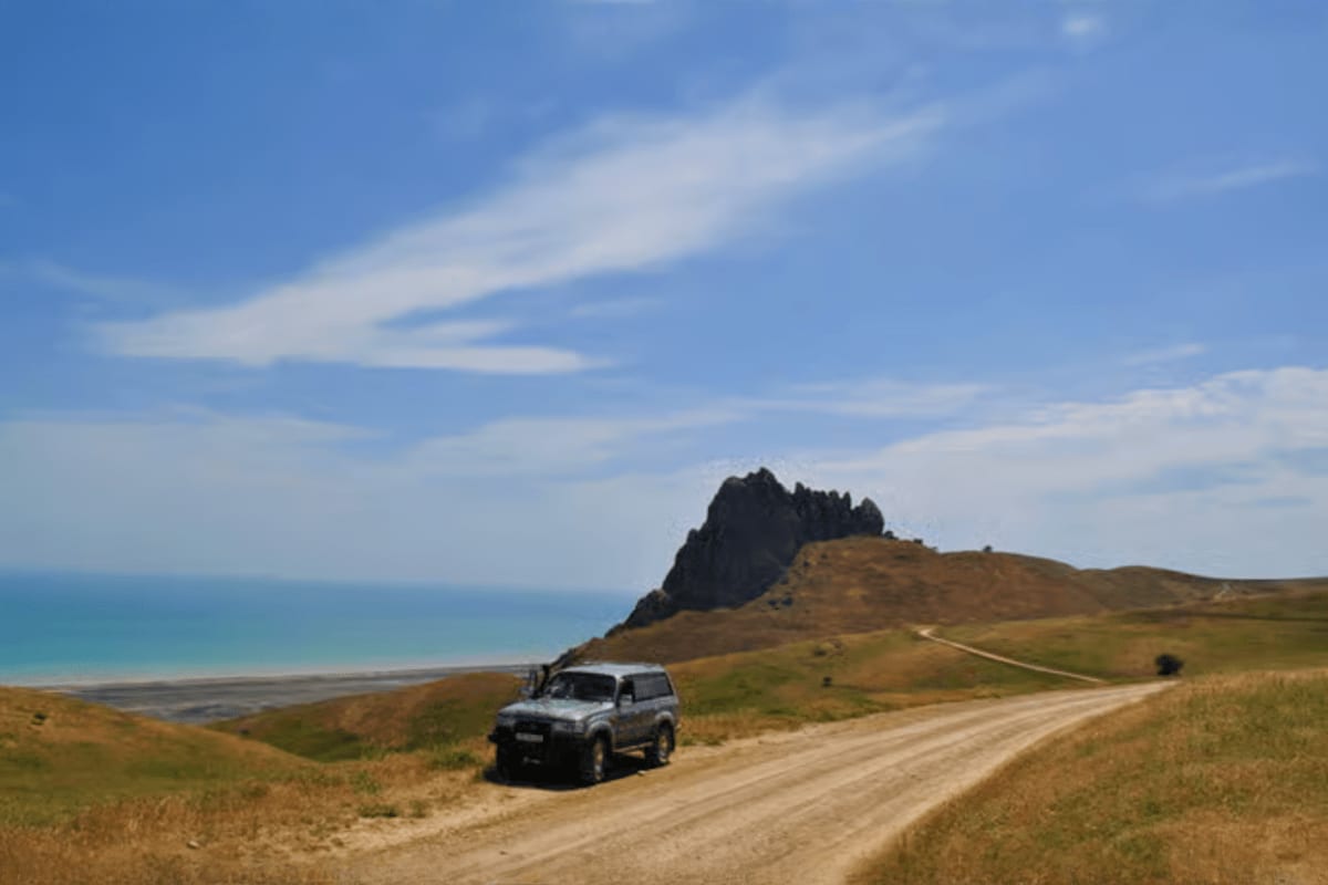 Baku to Shahdag Tour White SUV on dirt road with rocky mountain peak and turquoise sea coastline in background under blue sky