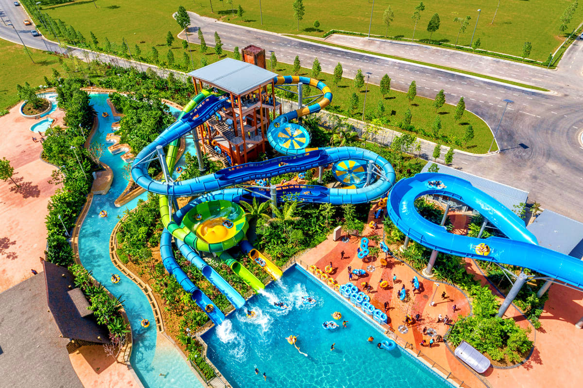 Aerial view of SplashMania Water Park with blue water slides, lazy river, swimming pools, and visitors on sandy beach areas s