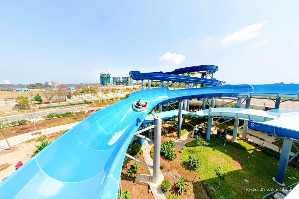Aerial view of SplashMania Water Park showing bright blue water slides with riders, tropical landscaping, and multiple slide