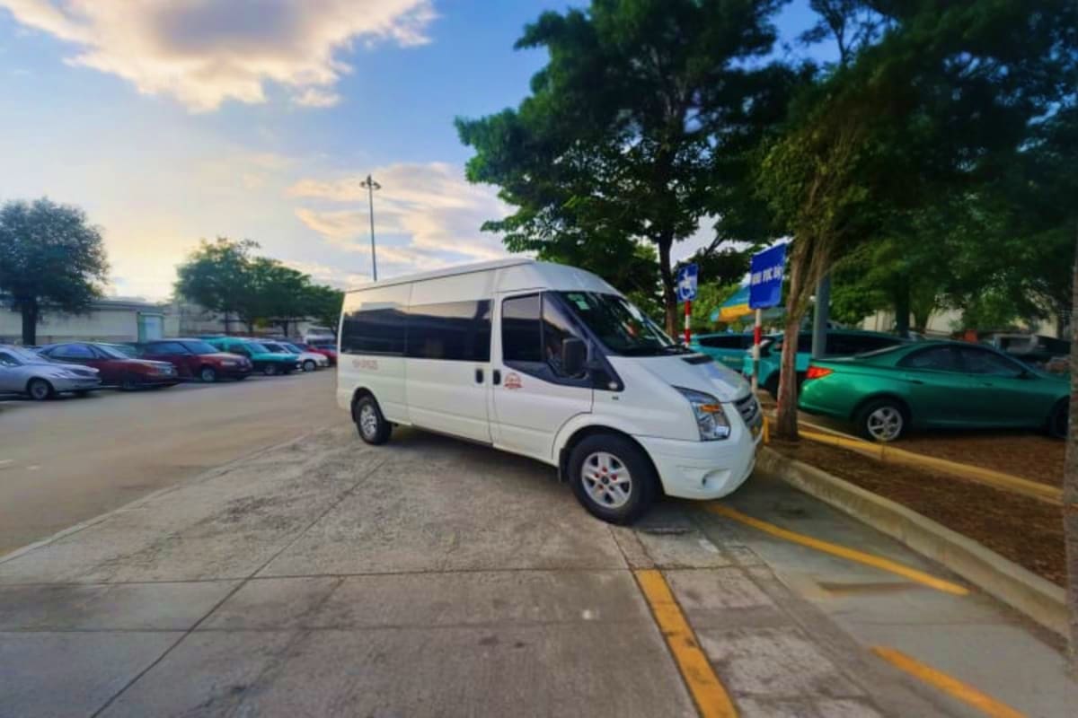 Ho Chi Minh Midnight Airport Pickup White passenger van parked at Ho Chi Minh City airport pickup area with parked cars and trees in background
