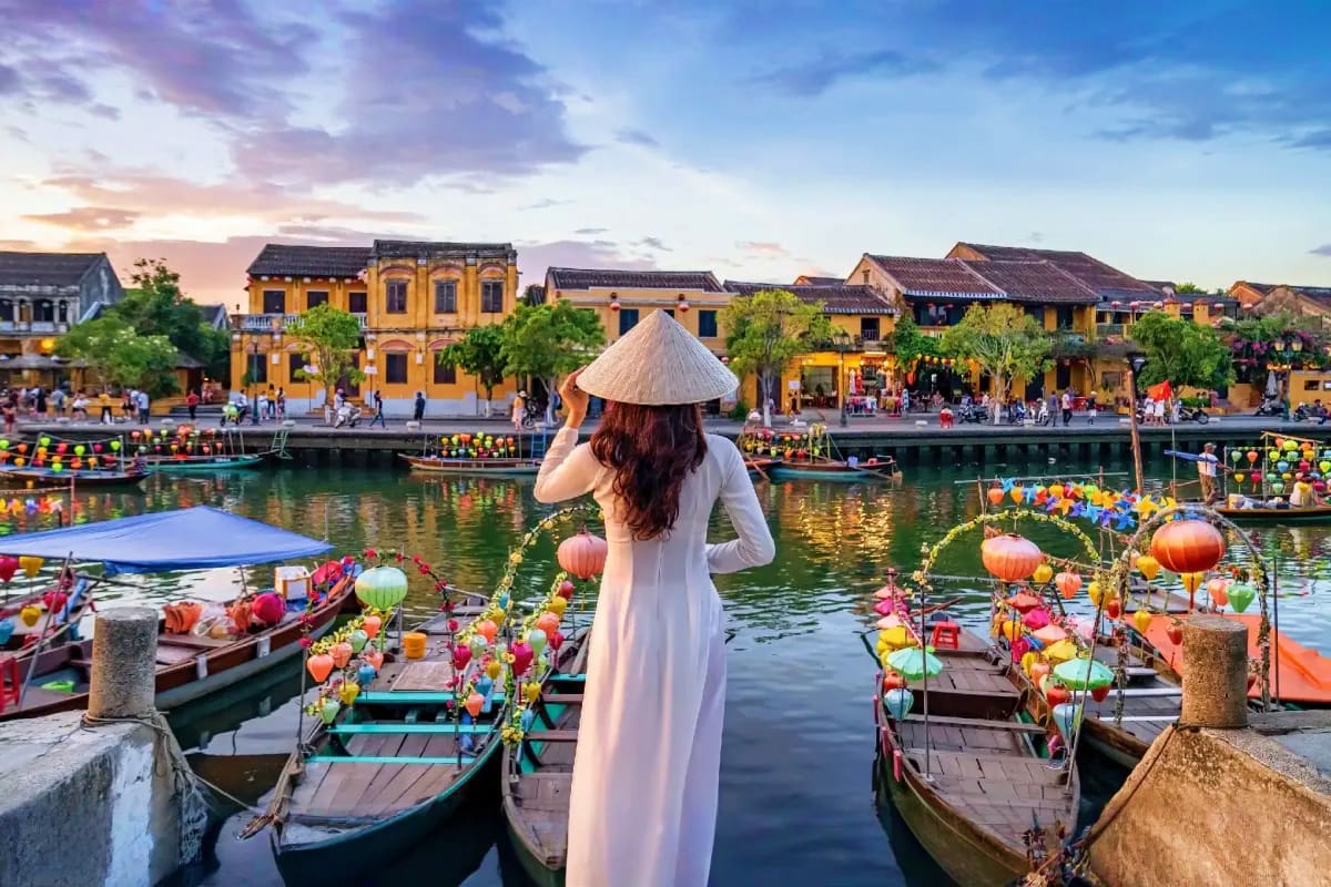 Cu Chi Tunnel And Full Day City Tour Woman in white áo dài and conical hat overlooking colorful lantern boats on Thu Bon River in Hoi An ancient town at sunset