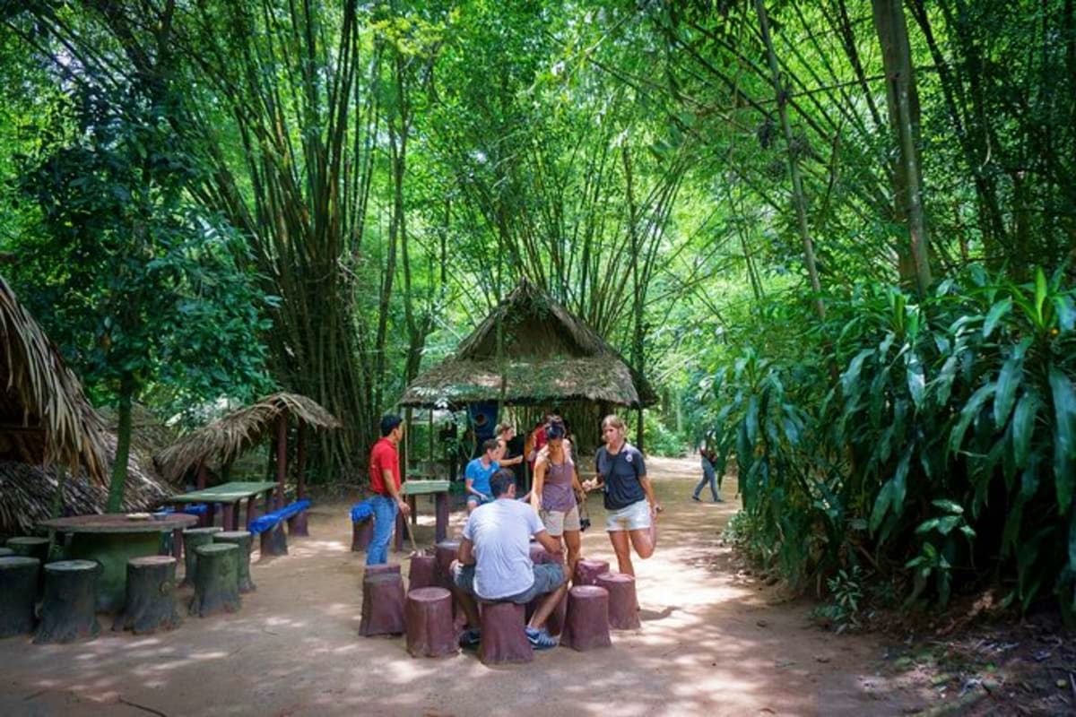 Tourists gathering at a thatched-roof shelter in a dense bamboo forest during a Cu Chi Tunnels tour activity