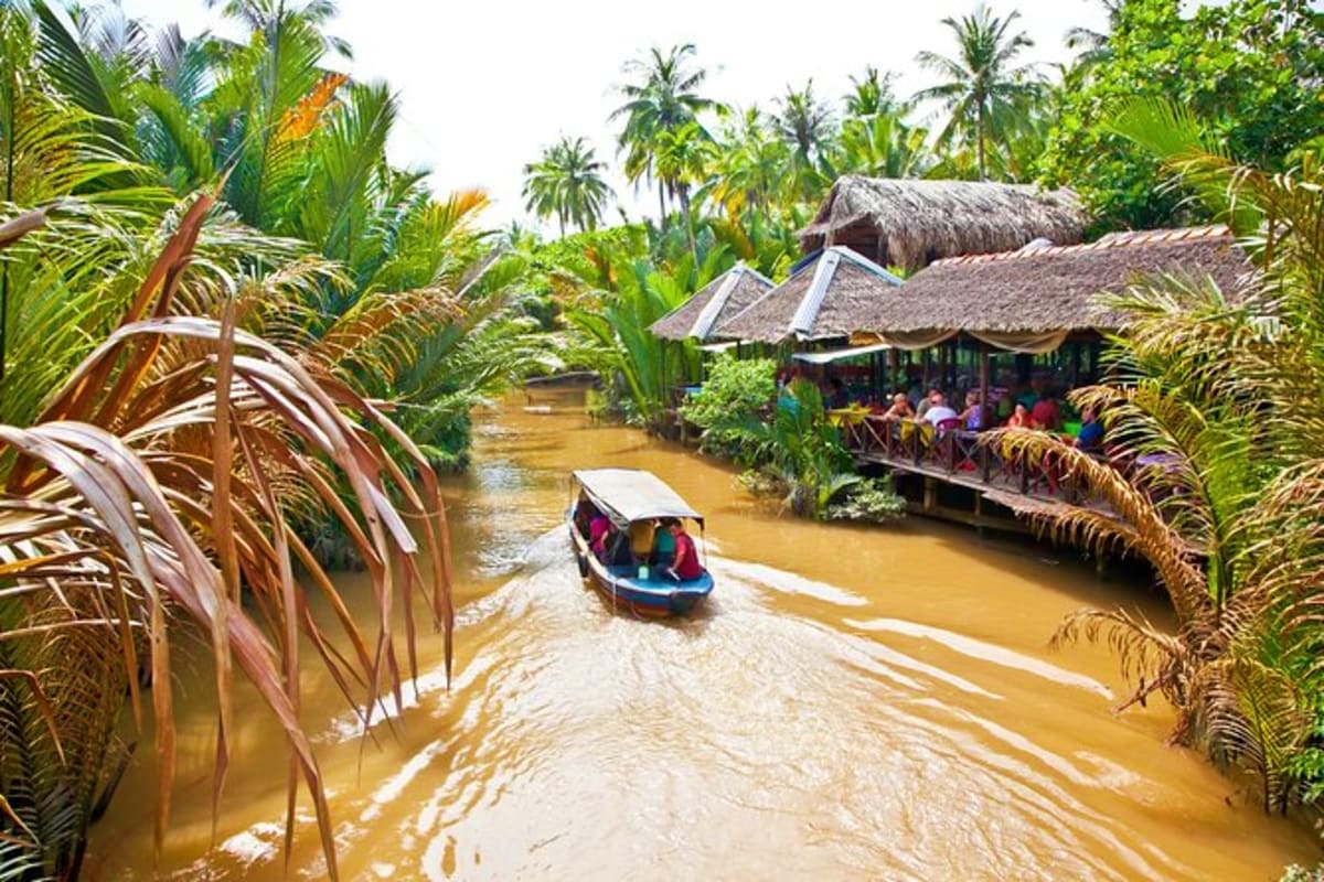 Cu Chi And Mekong Full Day Tour Small boat with tourists navigating through narrow waterway lined with palm trees and traditional thatched-roof houses in Mek