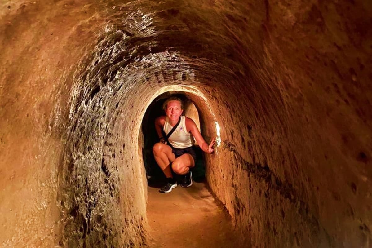 Cu Chi And Mekong Full Day Tour Woman crouching inside a narrow underground tunnel at Cu Chi Tunnels, holding a flashlight to illuminate the historic wartime