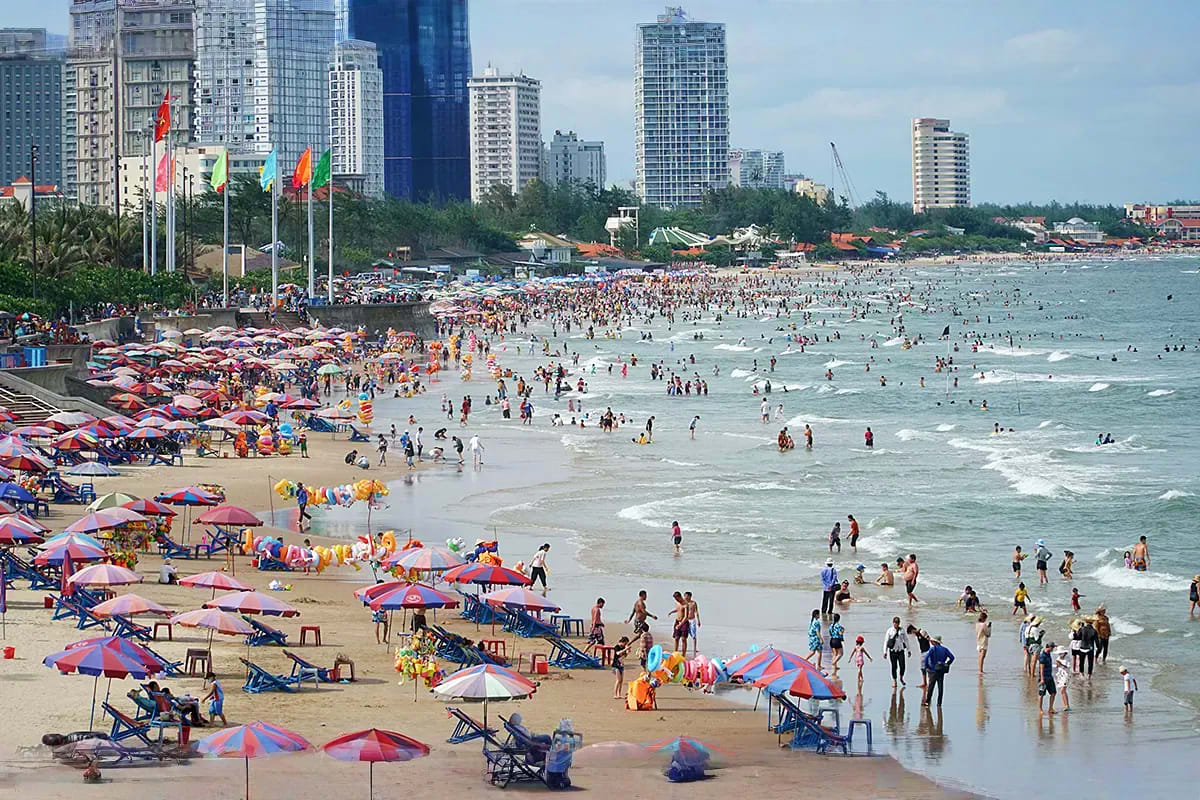 Vung Tau Day Trip Crowded Vung Tau beach with colorful umbrellas, swimmers in the water, and high-rise buildings in the background