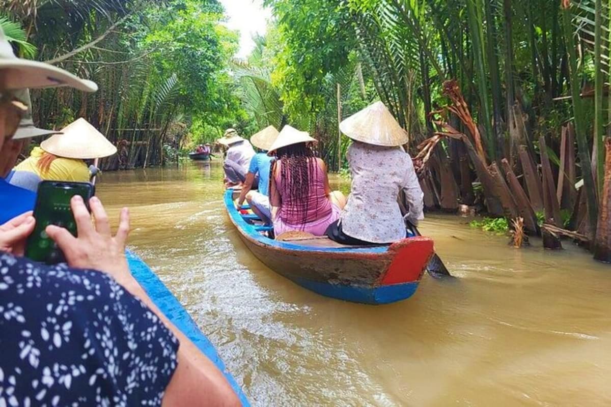 Mekong - Cai Be Tan Phong Island Sharing Tour Tourists in traditional Vietnamese conical hats riding wooden boats through narrow waterways surrounded by palm trees in the