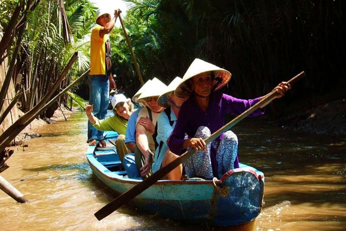 Mekong - Cai Be Tan Phong Island Sharing Tour Tourists paddling wooden boats through narrow waterways surrounded by tropical palm trees on Mekong Delta tour in Vietnam