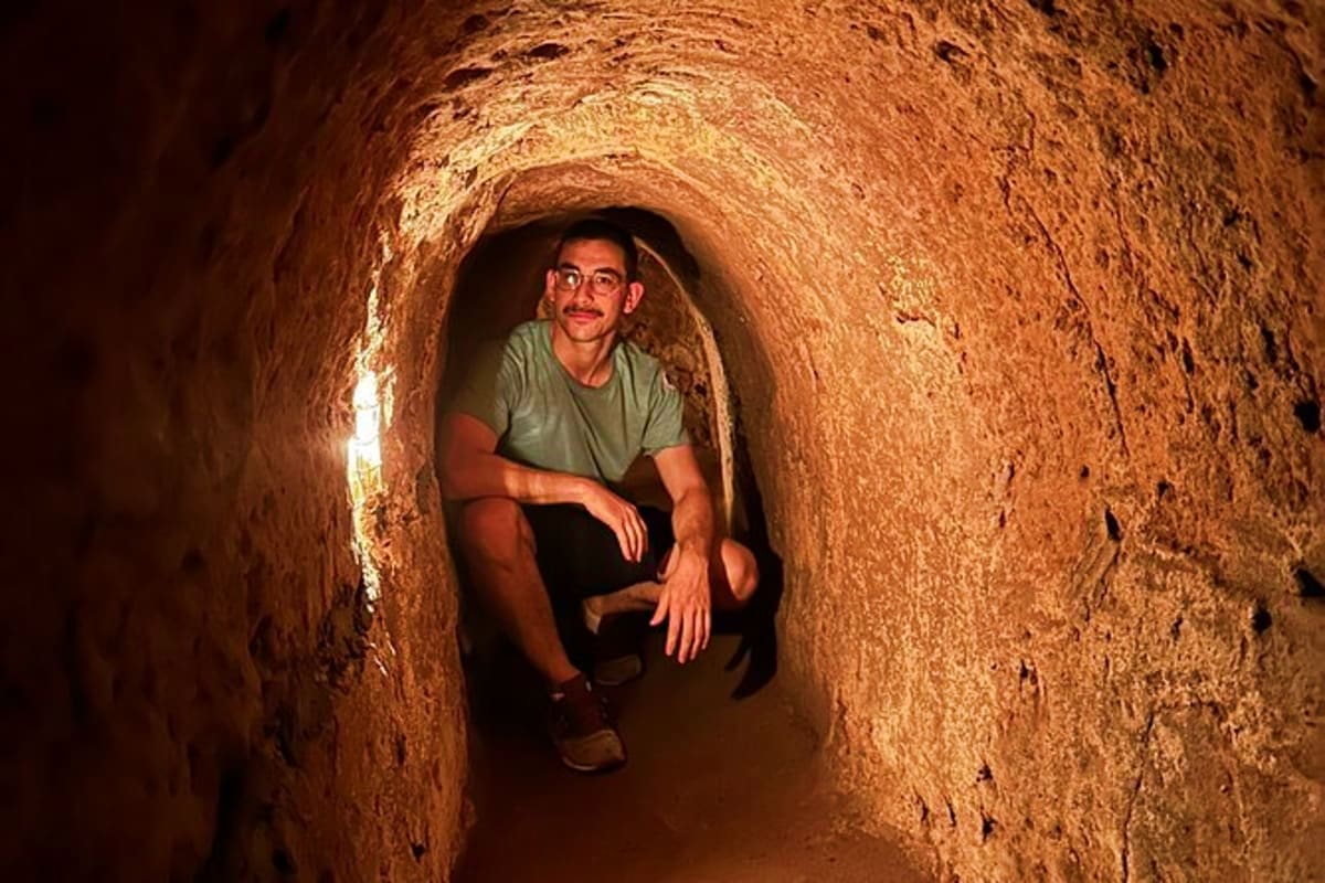 Half Day Cu Chi Tunnel Tour Man crouching inside narrow underground tunnel with brown earthen walls at Cu Chi Tunnels historical site