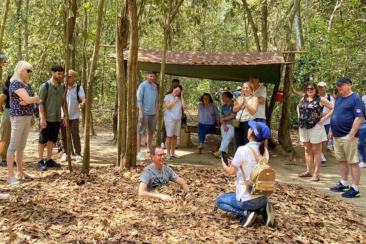 Group of tourists gathered at Cu Chi Tunnels historic site with guide demonstrating tunnel entrance in forested area