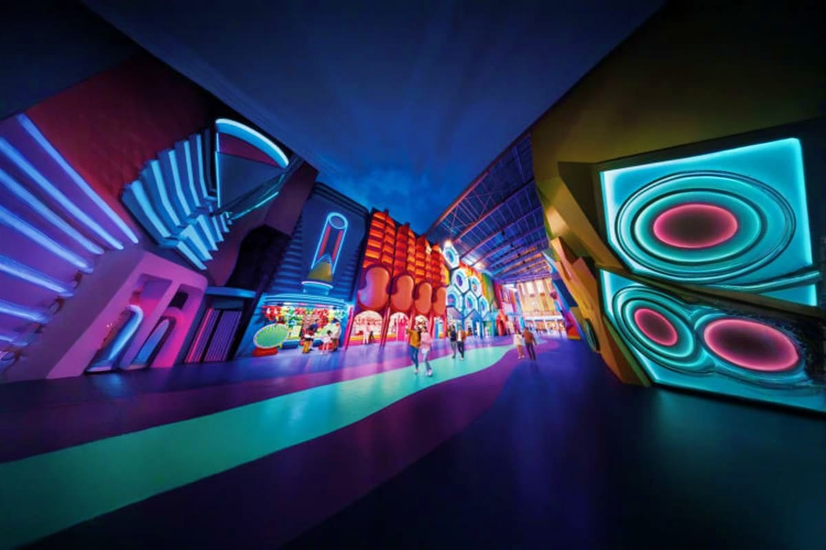 Vibrant neon-lit indoor theme park corridor with colorful digital displays, glowing signage, and visitors walking through fut