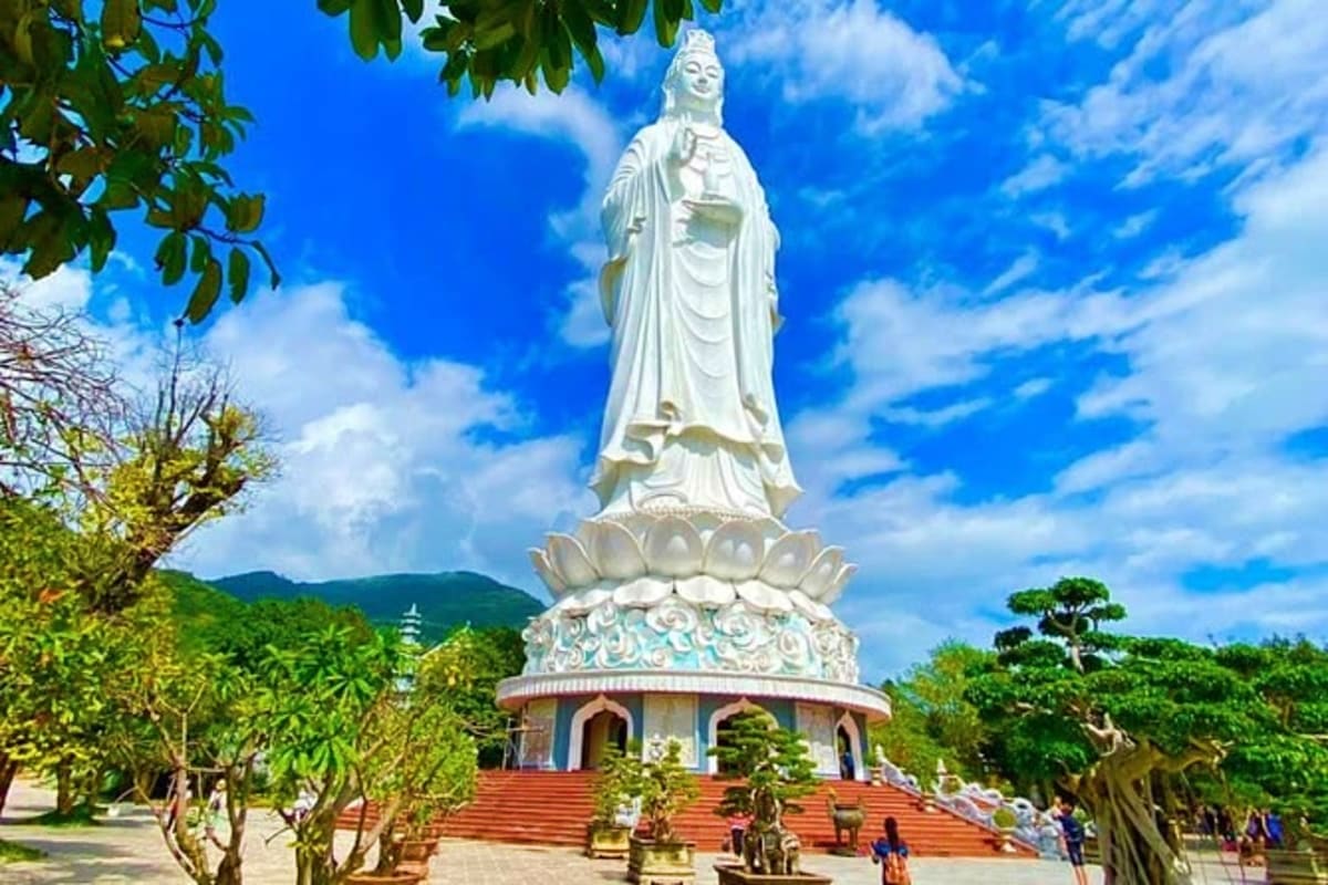 White marble statue of Guanyin bodhisattva on lotus pedestal at Linh Ung Pagoda with tropical mountains and visitors below