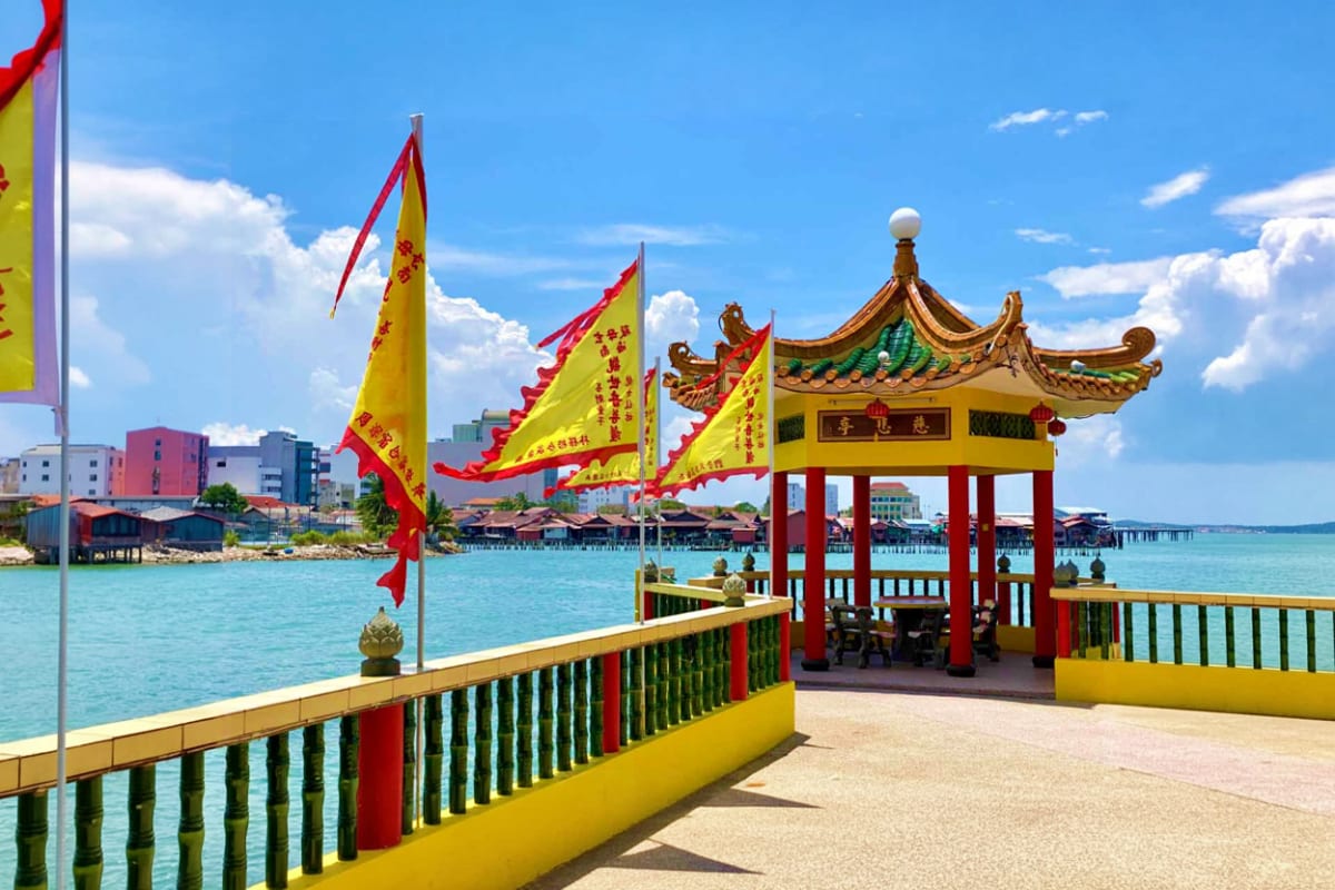 Chinese temple pavilion with red and yellow roof on wooden pier overlooking turquoise water in Penang, Malaysia
