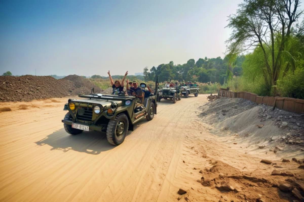 Tourists on a vintage military jeep driving down a dusty sand road with other jeeps behind them, passing through a rural land