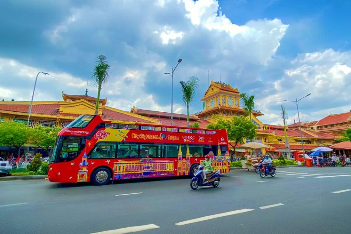 Red double-decker hop-on hop-off tour bus with colorful decorations driving past yellow temple buildings in Ho Chi Minh City