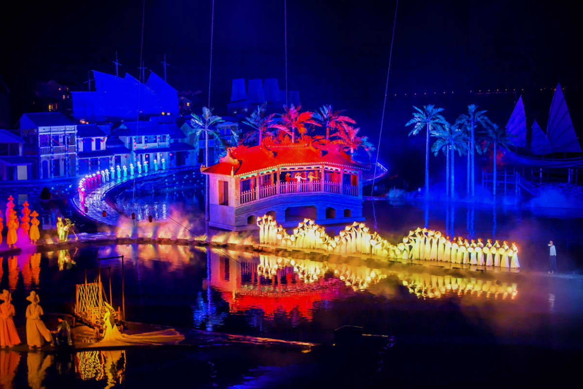 Hoi An Memories Show with Hoi An Impression Theme Park Illuminated traditional Vietnamese architecture reflected in water at night, featuring a red-roofed pavilion and blue-lit bui