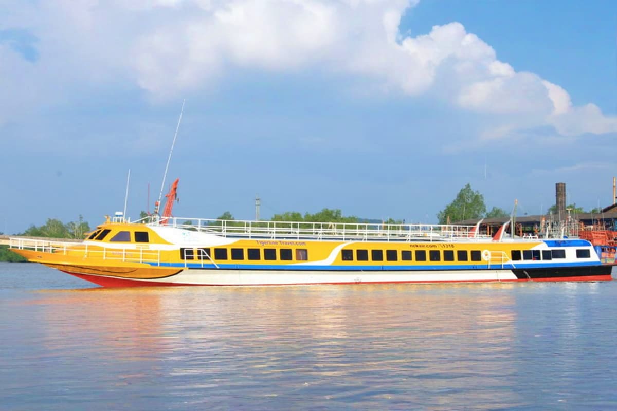 Yellow and blue high-speed ferry boat on calm water with industrial port buildings and trees in background under blue sky