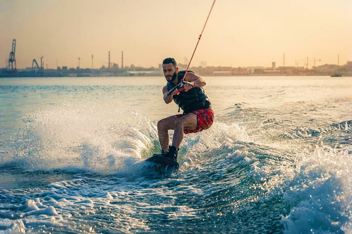 Man wakeboarding in ocean waves at sunset with industrial port and city skyline in background