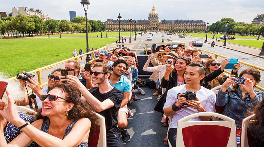 Tourists on open-top yellow Big Bus tour bus in Paris with Les Invalides dome and green park visible in background