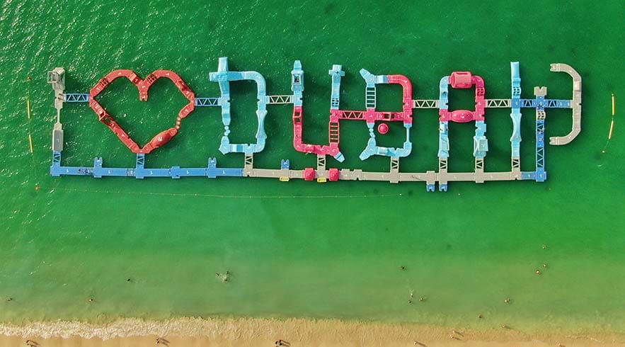 Aqua Fun Dubai Aerial view of Aqua Fun Dubai water park with colorful inflatable obstacle course modules arranged in ocean water near beach