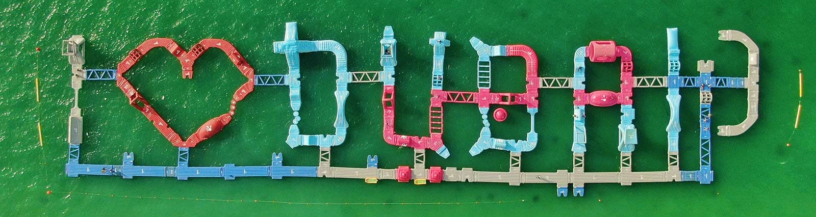 Aerial view of Aqua Fun Dubai water park with colorful inflatable obstacles and a red heart shape attraction on green water