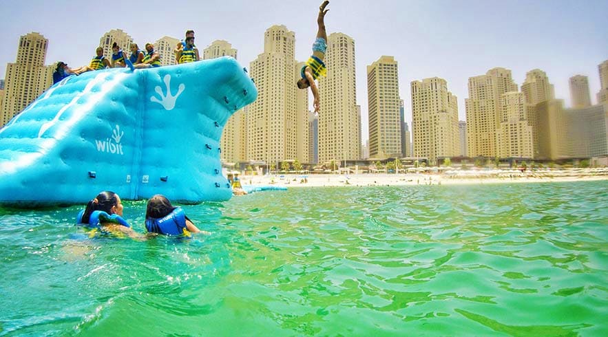 Aqua Fun Dubai People enjoying inflatable water slide activities in turquoise Gulf waters with Dubai Marina high-rise buildings in backgroun
