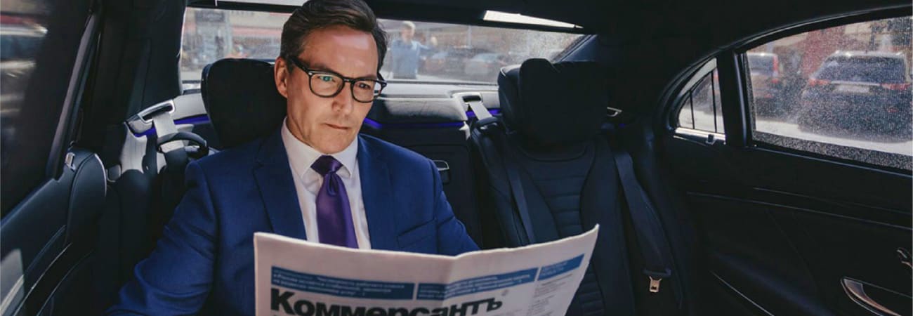 Businessman in blue suit and glasses reading newspaper in car backseat during airport transfer service