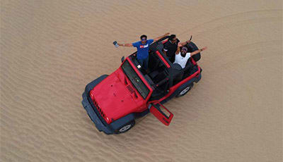 Dune Bashing Safari Jeddah Aerial view of red SUV driving across sandy desert with passengers enjoying a dune bashing safari experience