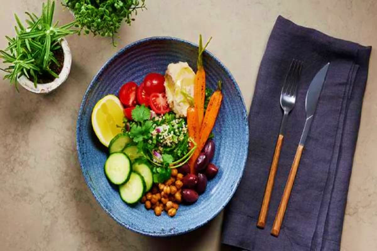 Colorful Buddha bowl with fresh vegetables, quinoa, and legumes on blue plate with fork, knife, and potted herbs on gray surf