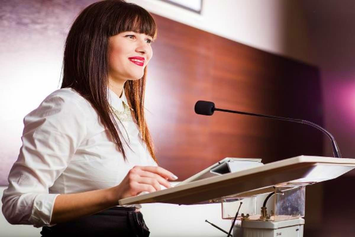 Professional woman in white blazer speaking at podium with microphone and papers during conference presentation