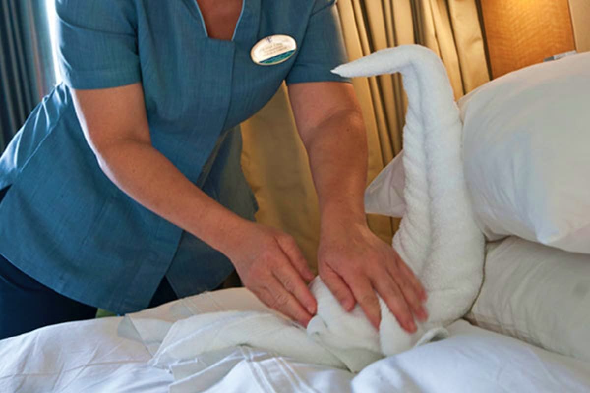 Hotel staff member in blue uniform making bed with white linens in cruise ship cabin