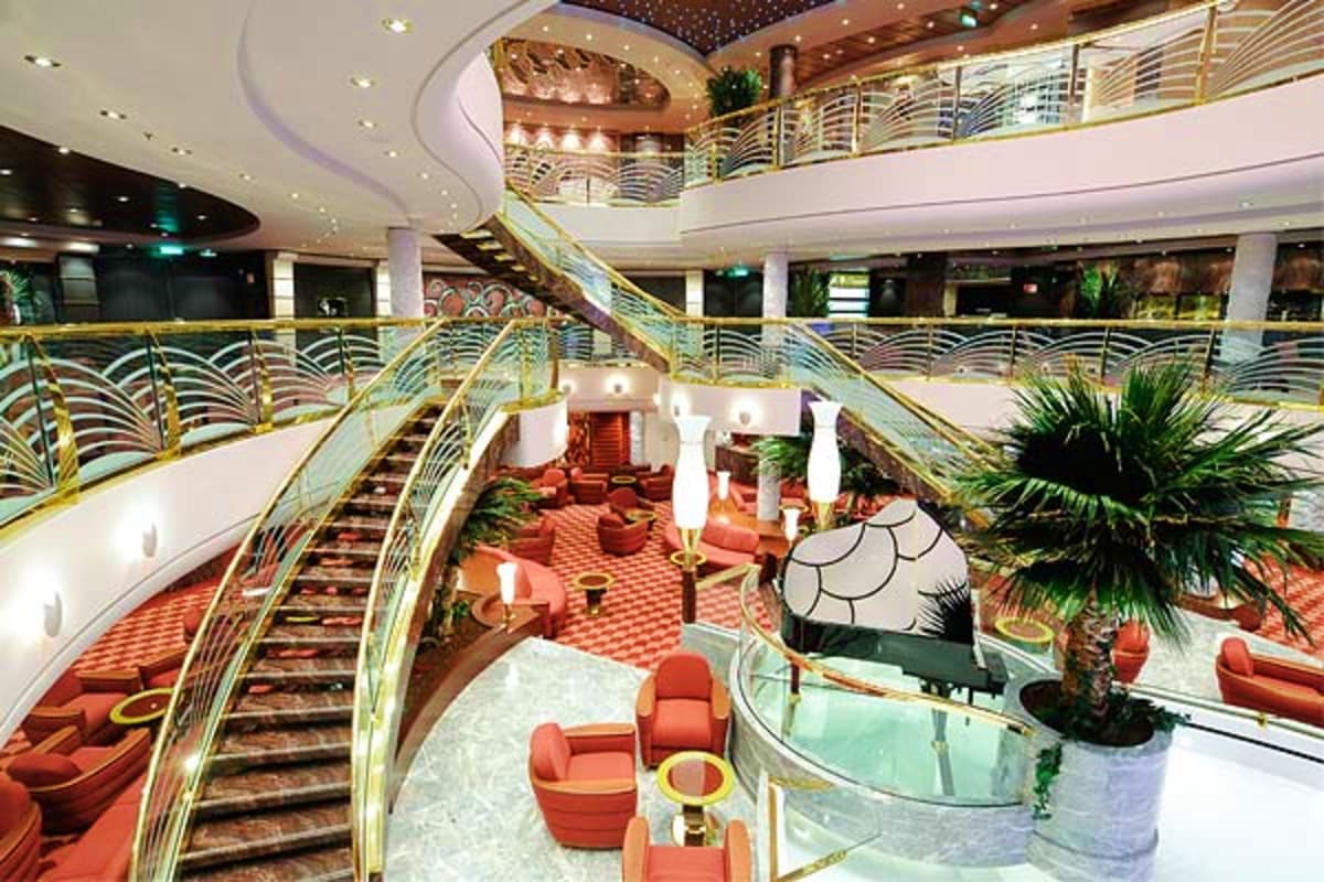 Luxury cruise ship interior atrium with multiple decks, grand staircase, gold railings, red carpet, palm plants, and modern l