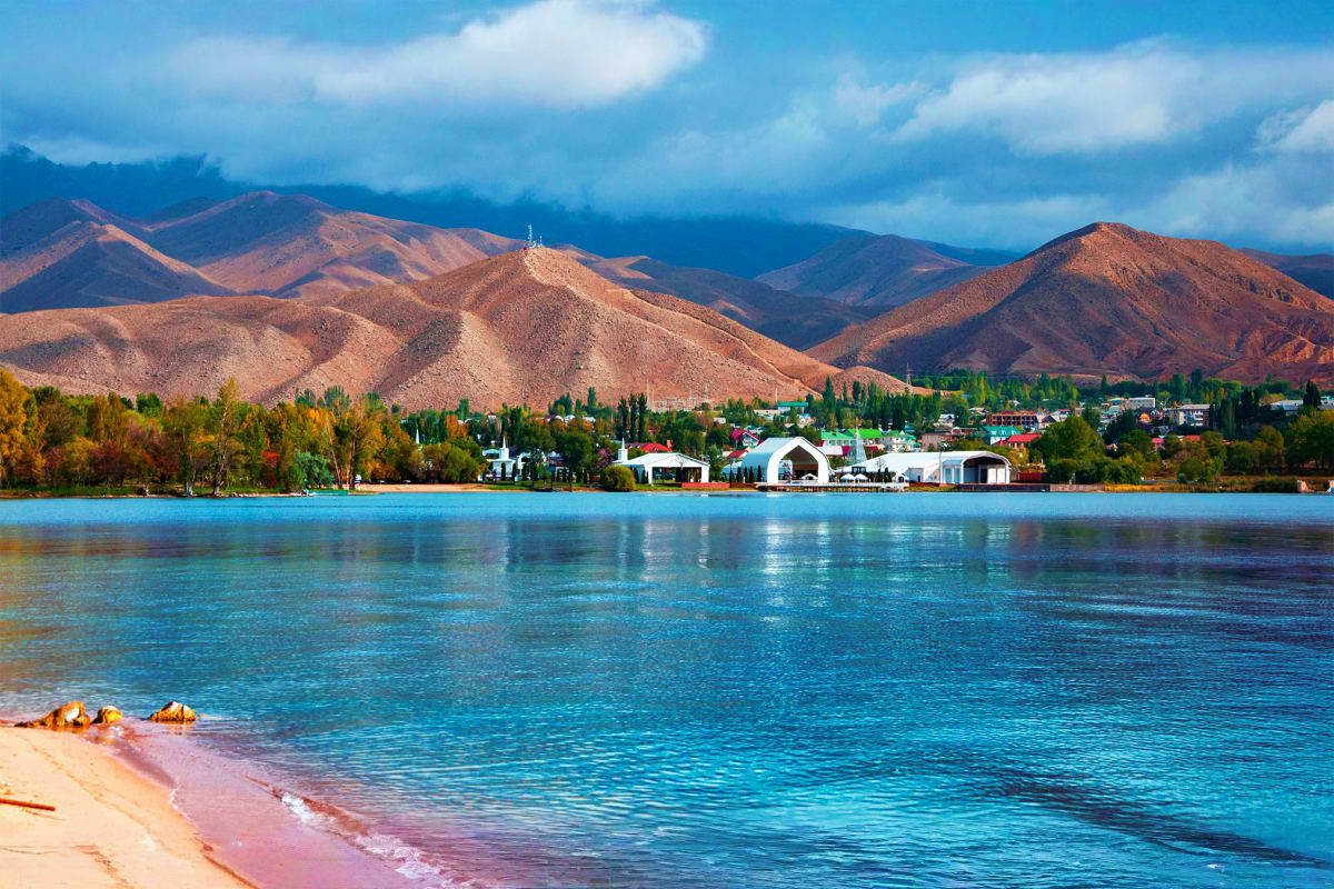 Scenic view of Issyk-Kul Lake with turquoise waters, sandy beach, tree-lined shore, residential buildings, and snow-capped mo